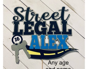 Puede incluir: Un adorno para pastel con las palabras "Street Legal" en blanco y negro, una llave con un diseño de carretera amarillo y negro, y el nombre "Alex" en azul y blanco. El número "16" está en la llave.