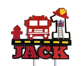 May include: Red, white, and black firefighter themed cake topper with a fire truck, fire hydrant, and a firefighter holding a ladder. The number one is on the firefighter's hat and the name "JACK" is at the bottom.