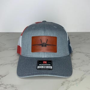 May include: A gray trucker hat with a brown leather patch featuring a jet plane design. The hat has a curved brim and a mesh back with red, white, and blue accents. The Richardson 112 Trucker hat is adjustable.