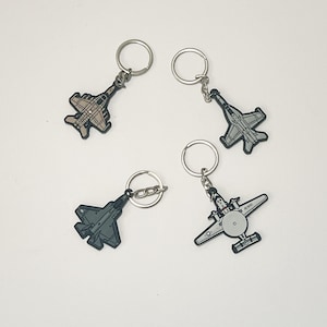 May include: Four airplane-shaped keychains. The keychains feature different aircraft designs in shades of gray, tan, and black. Each keychain has a silver metal ring for attaching keys.
