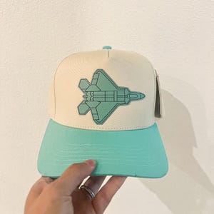 May include: A cream-colored baseball cap with a teal brim and a teal jet graphic. The cap has a flat brim and a snapback closure. The jet graphic is a detailed outline of a fighter jet.