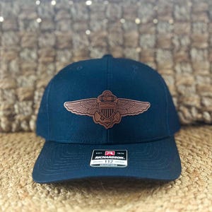 May include: A dark blue baseball cap with a brown leather patch featuring a winged anchor and the text "U.S. NAVY". The cap has a black adjustable strap with the text "Richardson 1970" and "112" printed on it.