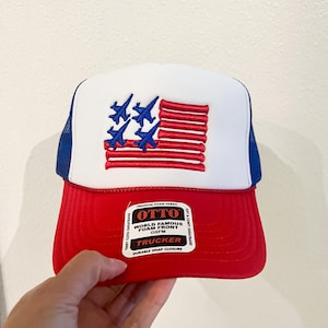 May include: A red, white, and blue trucker hat with an embroidered American flag design. The hat features a red brim, a white front panel with the flag, and a blue mesh back. The hat has a snap closure.