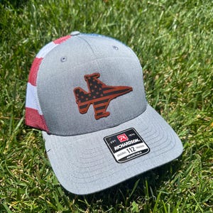May include: Gray Richardson 112 trucker hat with a brown leather patch in the shape of a jet, filled with a red and white star pattern. The back of the hat features a red, white, and blue striped design. The hat is sitting on green grass.
