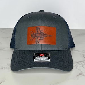 May include: A gray trucker hat with a navy blue mesh back and brim. The hat features a brown leather patch with a detailed outline of a jet. The patch is rectangular. The hat has the text "RICHARDSON" and "ORIGINAL 112 TRUCKER" on the front.