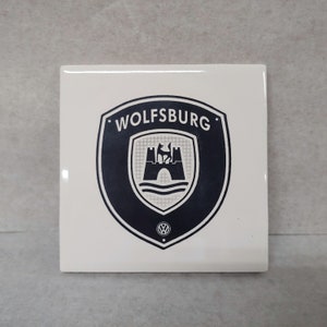 May include: White ceramic tile with a black and white Volkswagen Wolfsburg logo. The logo features a castle with a wolf on top, a river, and the text "WOLFSBURG".
