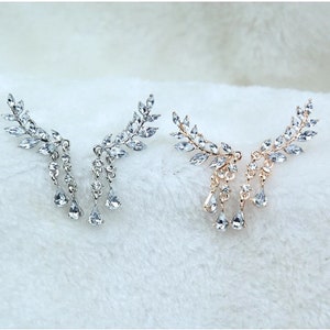 May include: Two ear cuff earrings, one silver-toned and one gold-toned, each featuring a leaf design with clear crystal accents. Dangling teardrop-shaped crystals add a touch of elegance. Perfect for special occasions.