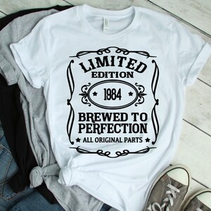May include: White t-shirt with a black graphic that reads "LIMITED EDITION 1984 BREWED TO PERFECTION ALL ORIGINAL PARTS." The shirt is folded with other shirts and jeans. Gray sneakers are visible.