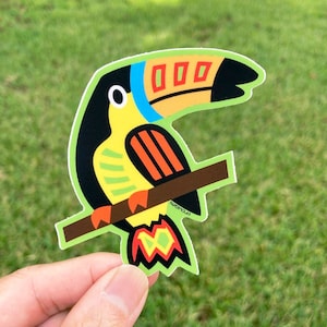 May include: A colorful toucan sticker with a black head, yellow body, orange and red accents, and a black beak with a yellow stripe. The toucan is perched on a brown branch.