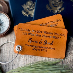 Puede incluir: Dos etiquetas de equipaje de cuero marrón con el texto "In Life, It's Not Where You Go, It's Who You Travel With" y "Where You Go, I With". La etiqueta inferior tiene los nombres "Rena & Brad" y la fecha "May 5, 2018".