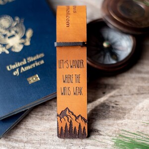 May include: A brown leather bookmark with the text "Let's wander where the wifi is weak" and a mountain range illustration. The bookmark is next to a blue passport and a compass.