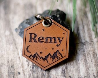 Peut inclure: Une médaille de chien hexagonale en cuir marron avec un motif de montagne et de lune et le nom "Remy" gravé dessus.