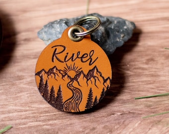Peut inclure: Une étiquette pour animaux de compagnie ronde en cuir bronzé avec un motif de montagne et de rivière. Le mot "River" est écrit en haut. Elle est munie d'un anneau métallique pour l'attacher. Le fond est une surface en bois.