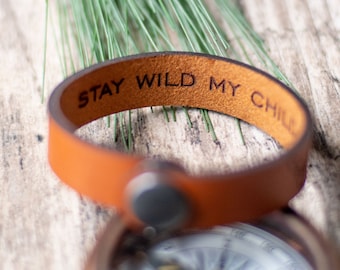 Puede incluir: Un brazalete de cuero marrón con el texto "STAY WILD MY CHILD" grabado en él. El brazalete está sobre una superficie de madera con una brújula en el fondo.
