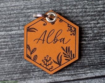 Puede incluir: Un llavero de cuero marrón con una etiqueta hexagonal. La etiqueta tiene un diseño floral y el nombre "Alba" grabado en cursiva.