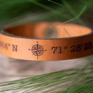 Op de afbeelding: Een bruin leren armband met een kompasontwerp en de coördinaten "71° 28' 25.4" erop gegraveerd.