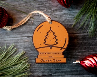 May include: A brown wooden Christmas ornament shaped like a snow globe with a small Christmas tree inside. The ornament has the text "Baby's First Christmas 2021 Oliver Sean" on the bottom.