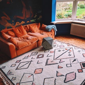 May include: A living room with a large orange couch, a geometric patterned rug, and a geometric cube ottoman. The room has a blue wall and a large window with a view of a garden.