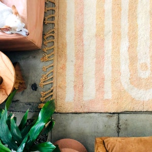 May include: A white and beige striped rug with fringe on the edges. The rug is on a concrete floor with a potted plant, a wooden stool, and a brown leather chair.