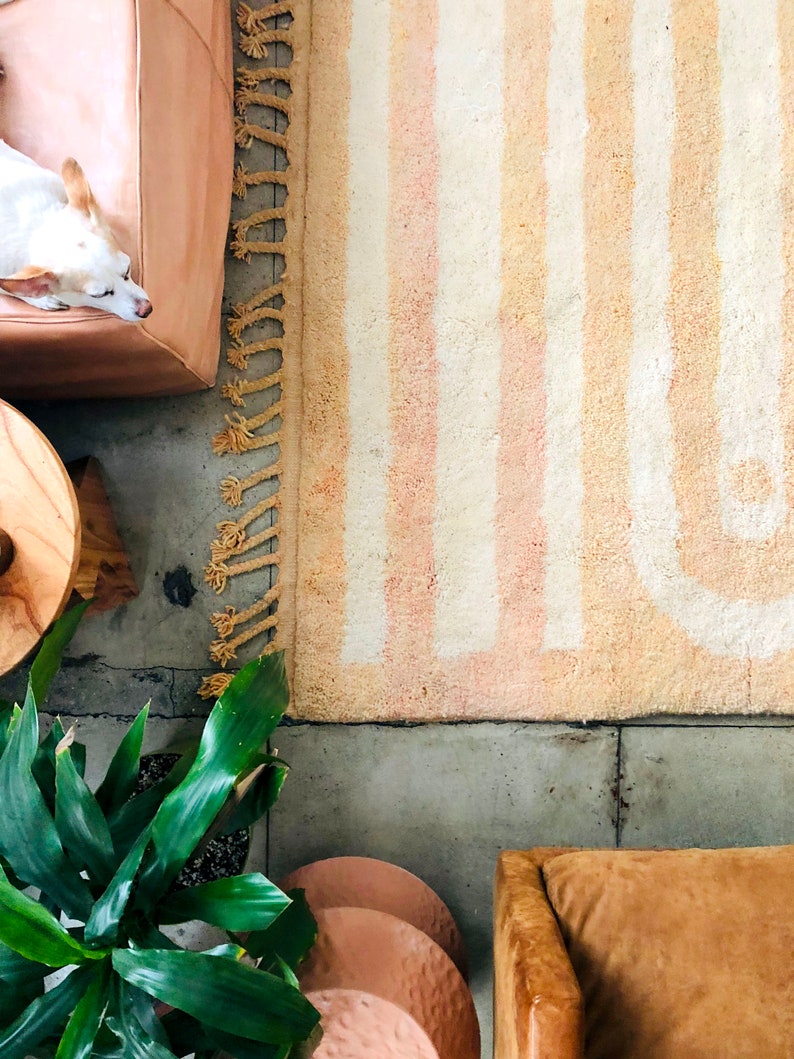 May include: A white and beige striped rug with fringe on the edges. The rug is on a concrete floor with a potted plant, a wooden stool, and a brown leather chair.