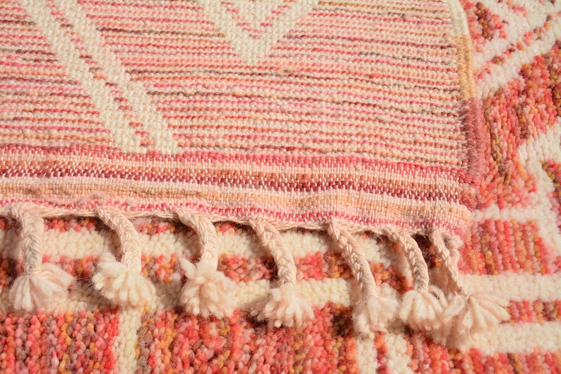 Puede incluir: Primer plano de una alfombra tejida rosa y blanca con flecos. La alfombra tiene un patr&oacute;n geom&eacute;trico y est&aacute; hecha de fibras suaves y esponjosas.