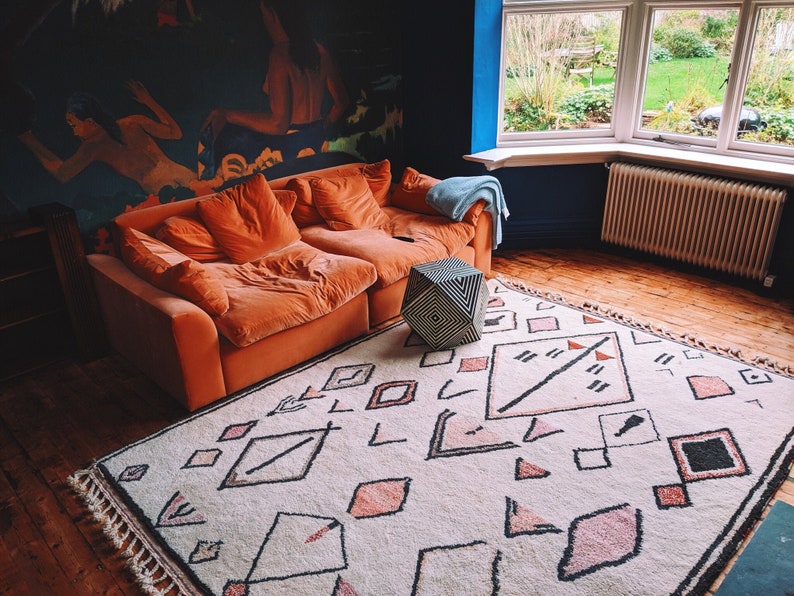 May include: A living room with a large orange couch, a geometric patterned rug, and a geometric cube ottoman. The room has a blue wall and a large window with a view of a garden.