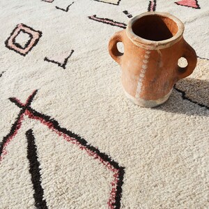 May include: A terracotta pot with two handles sits on a white rug with a geometric pattern. The rug has black and red lines.