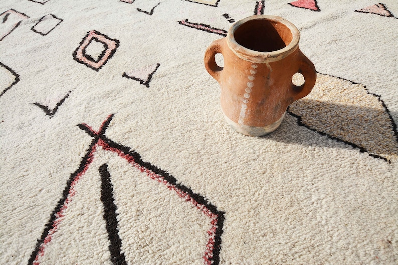 May include: A terracotta pot with two handles sits on a white rug with a geometric pattern. The rug has black and red lines.