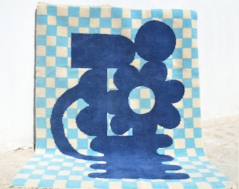 Puede incluir: Una alfombra a cuadros azules y blancos con un gran diseño de flor abstracta azul. La flor tiene un círculo grande en el centro y varios pétalos redondeados.