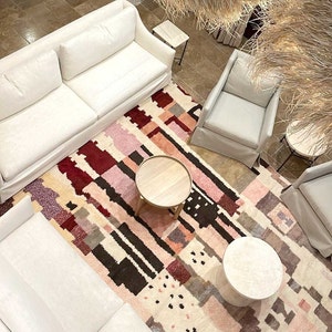 Puede incluir: Una alfombra blanca y mullida con un patr&oacute;n geom&eacute;trico en tonos rosa, rojo y marr&oacute;n. Dos sof&aacute;s blancos y dos sillas blancas est&aacute;n dispuestos alrededor de la alfombra, con dos mesas de caf&eacute; de madera y una mesa de caf&eacute; blanca.