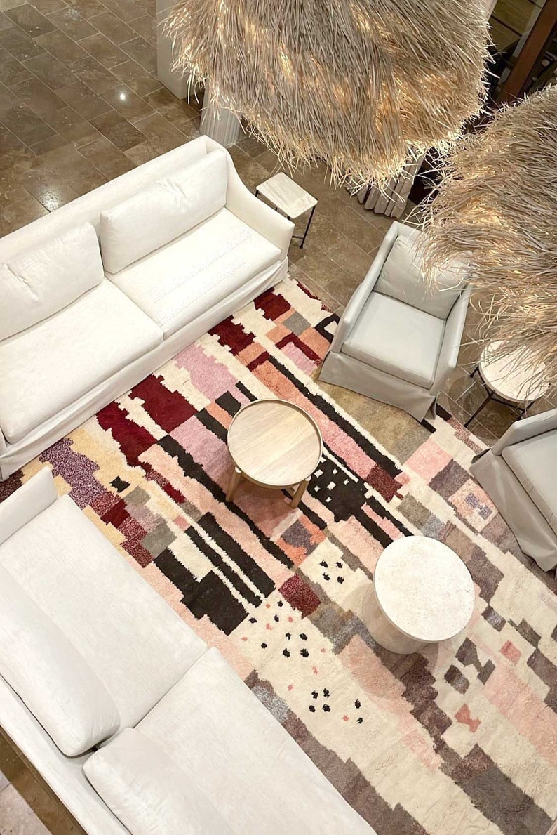 Puede incluir: Una alfombra blanca y mullida con un patr&oacute;n geom&eacute;trico en tonos rosa, rojo y marr&oacute;n. Dos sof&aacute;s blancos y dos sillas blancas est&aacute;n dispuestos alrededor de la alfombra, con dos mesas de caf&eacute; de madera y una mesa de caf&eacute; blanca.