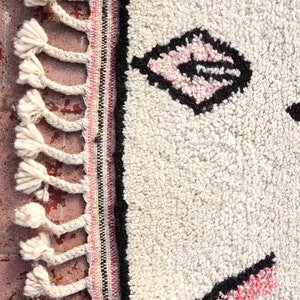 May include: Close-up of a white rug with black and pink geometric designs. The rug has a fringe border with white tassels.