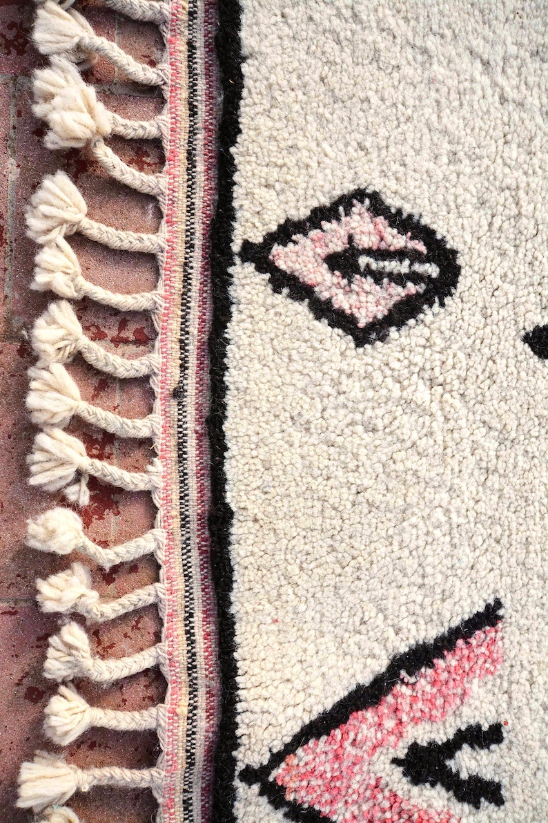 May include: Close-up of a white rug with black and pink geometric designs. The rug has a fringe border with white tassels.
