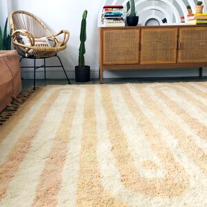 May include: A light pink and white striped rug with a curved pattern. The rug is in a living room with a wicker chair, a wooden cabinet, and a cactus plant.