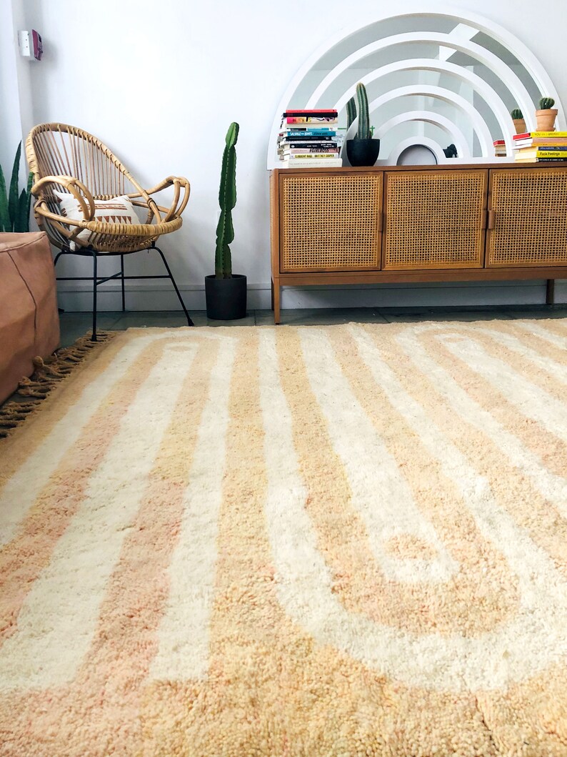 May include: A light pink and white striped rug with a curved pattern. The rug is in a living room with a wicker chair, a wooden cabinet, and a cactus plant.
