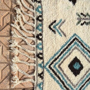 May include: A white wool rug with a geometric pattern in shades of blue, black, and gray. The rug has fringe on all sides.