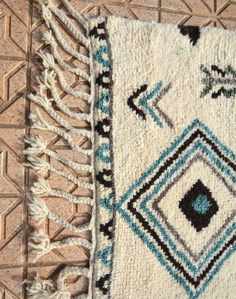 May include: A white wool rug with a geometric pattern in shades of blue, black, and gray. The rug has fringe on all sides.