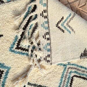 May include: A white rug with a geometric pattern in shades of blue, brown, and gray. The rug has fringe on the edges.