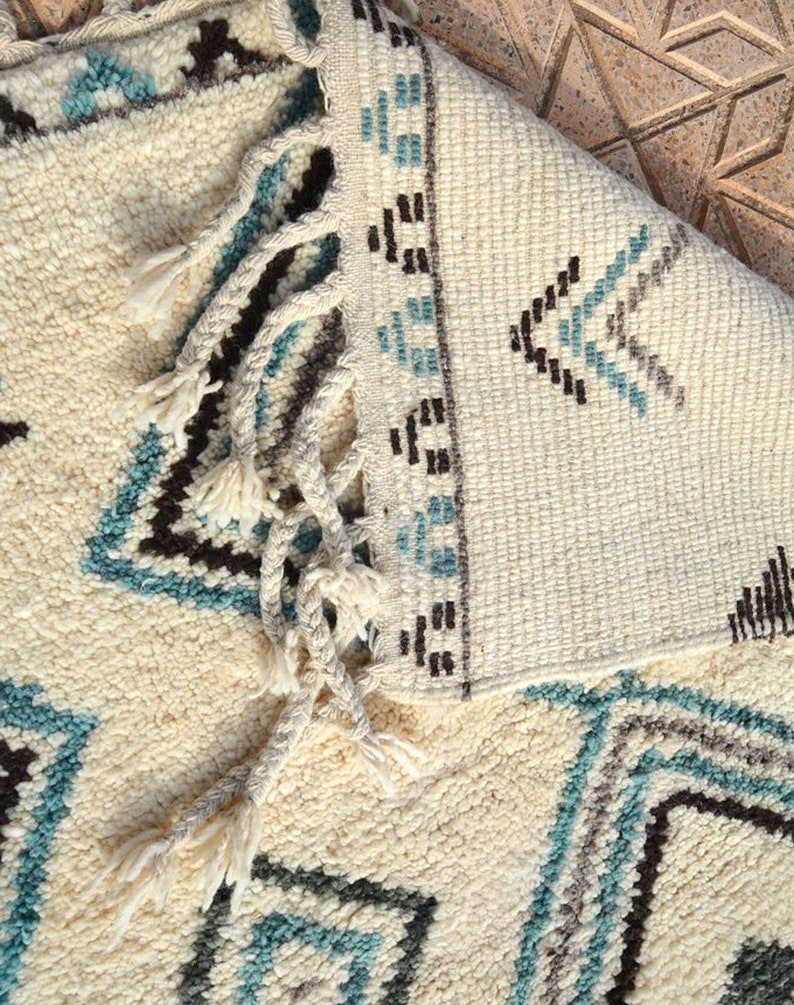 May include: A white rug with a geometric pattern in shades of blue, brown, and gray. The rug has fringe on the edges.
