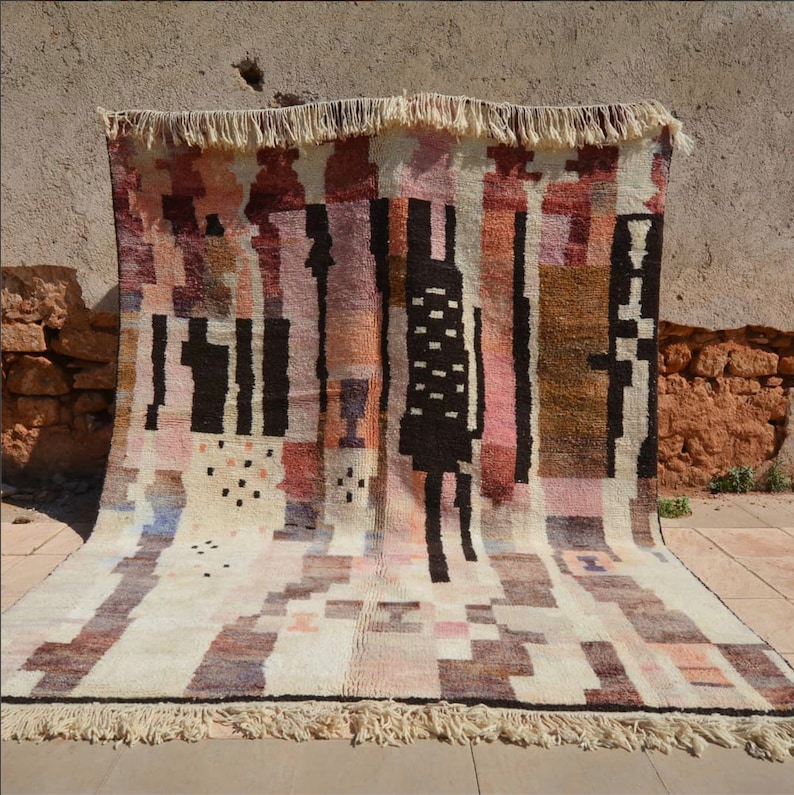 Puede incluir: Una alfombra grande y rectangular con un patr&oacute;n geom&eacute;trico en tonos marrones, rosas, blancos y negros. La alfombra tiene un borde de flecos.
