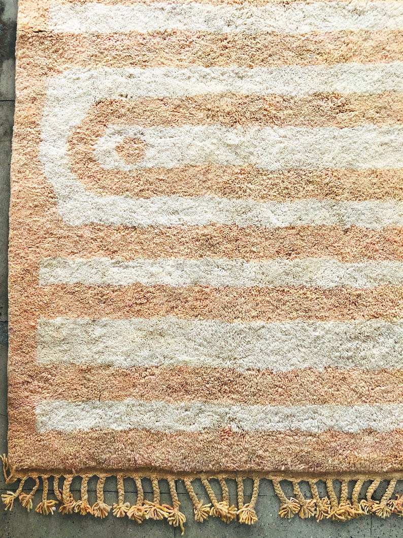 May include: A beige and white striped rug with a large, abstract, curved design in the center. The rug has a fringe border.