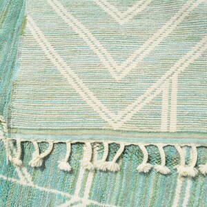 May include: A light blue and white woven rug with a geometric pattern and fringe. The rug has a subtle green hue throughout.