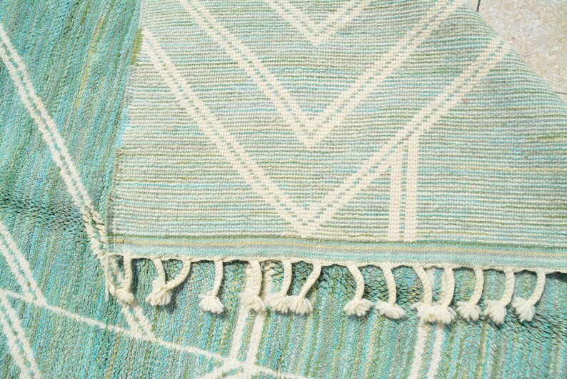 May include: A light blue and white woven rug with a geometric pattern and fringe. The rug has a subtle green hue throughout.
