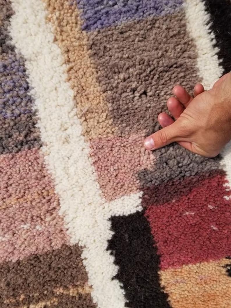 Puede incluir: Primer plano de una alfombra colorida y multitexturada con una mano descansando sobre la superficie. La alfombra presenta una variedad de colores, incluyendo rosa, marr&oacute;n, blanco y negro.