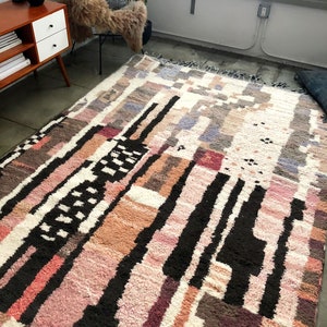 Puede incluir: Una alfombra colorida con un patr&oacute;n abstracto, fondo blanco y formas geom&eacute;tricas negras, rosas, marrones y grises. La alfombra tiene un borde de flecos.