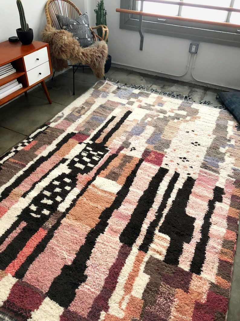 Puede incluir: Una alfombra colorida con un patr&oacute;n abstracto, fondo blanco y formas geom&eacute;tricas negras, rosas, marrones y grises. La alfombra tiene un borde de flecos.