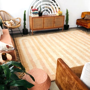May include: A living room with a light brown rug with white stripes, a brown leather couch, a wicker chair, a wooden cabinet with a white arched mirror above it, and a potted cactus plant.