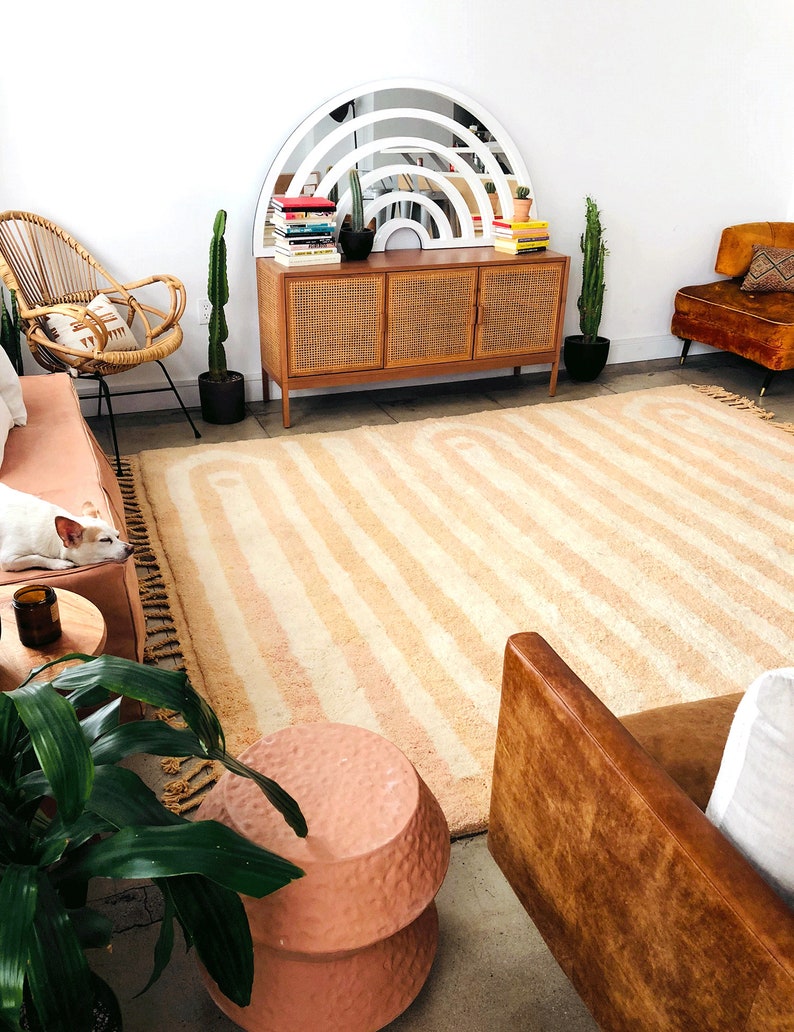May include: A living room with a light brown rug with white stripes, a brown leather couch, a wicker chair, a wooden cabinet with a white arched mirror above it, and a potted cactus plant.