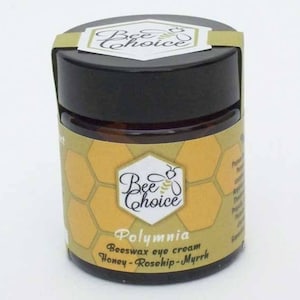 Puede incluir: Un pequeño frasco redondo de crema para ojos Bee Choice Polymnia Beeswax. El frasco tiene una tapa negra y una etiqueta con un patrón de panal y las palabras "Bee Choice" y "Polymnia Beeswax eye cream Honey-Rosehip-Myrrh."