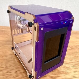 May include: A compact computer case with a purple top and front. It features clear acrylic sides, a small screen, and a fan. The frame is constructed from wood and secured with black screws. The case is designed for a small form factor.
